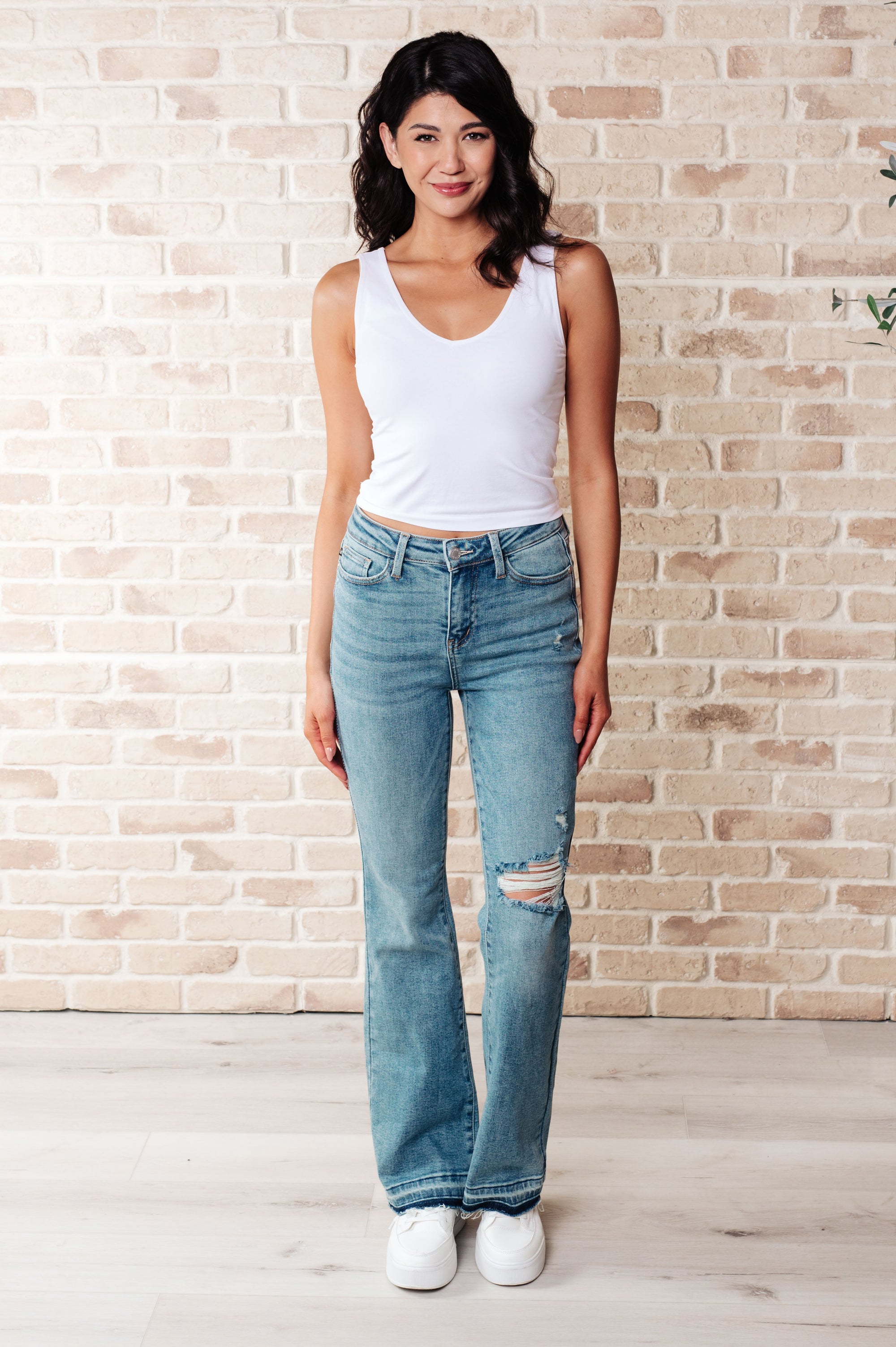 Woman wearing a white tank top and blue jeans standing against a brick wall.