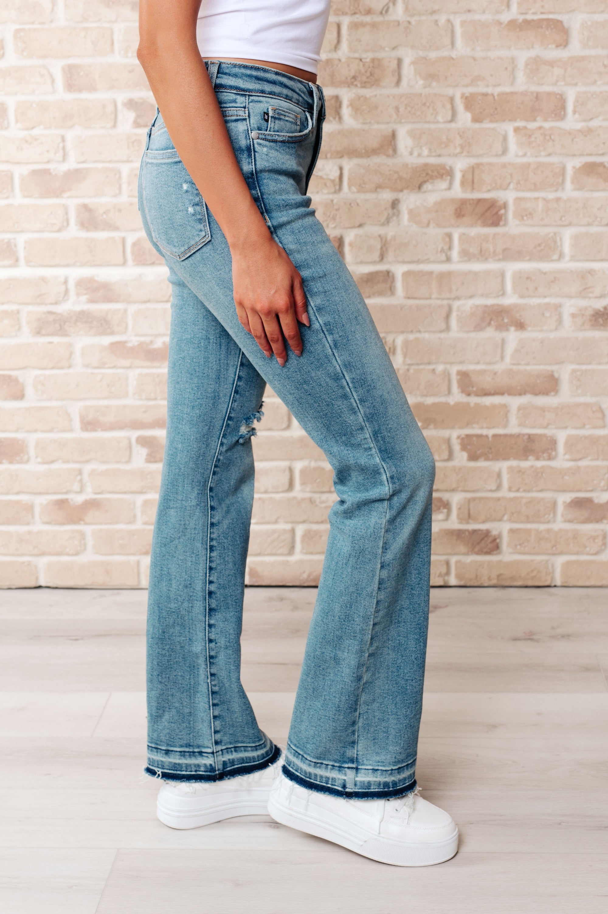 Person wearing light blue jeans and white sneakers against a brick wall.
