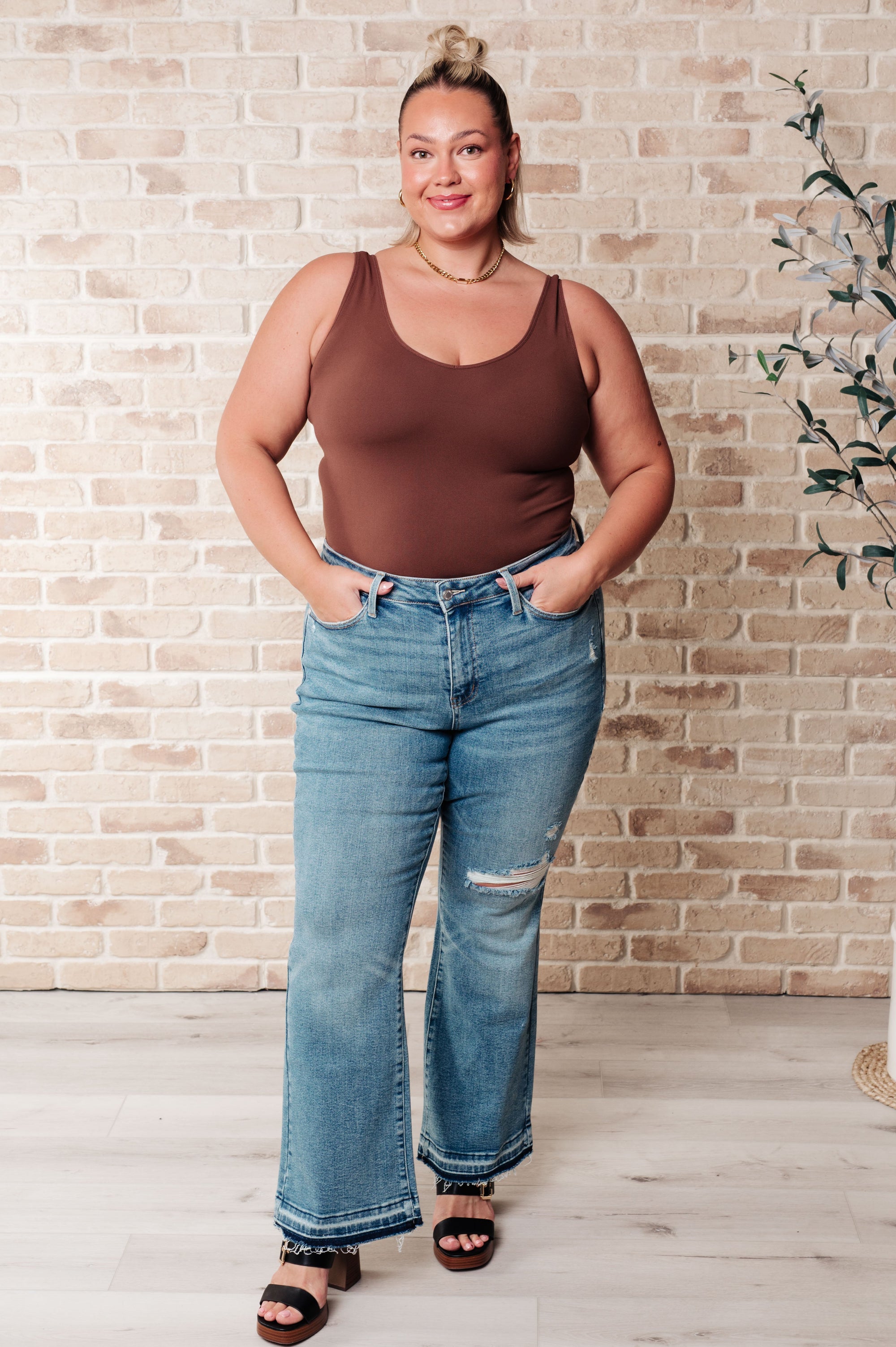 Woman wearing a brown tank top and blue jeans standing against a brick wall.