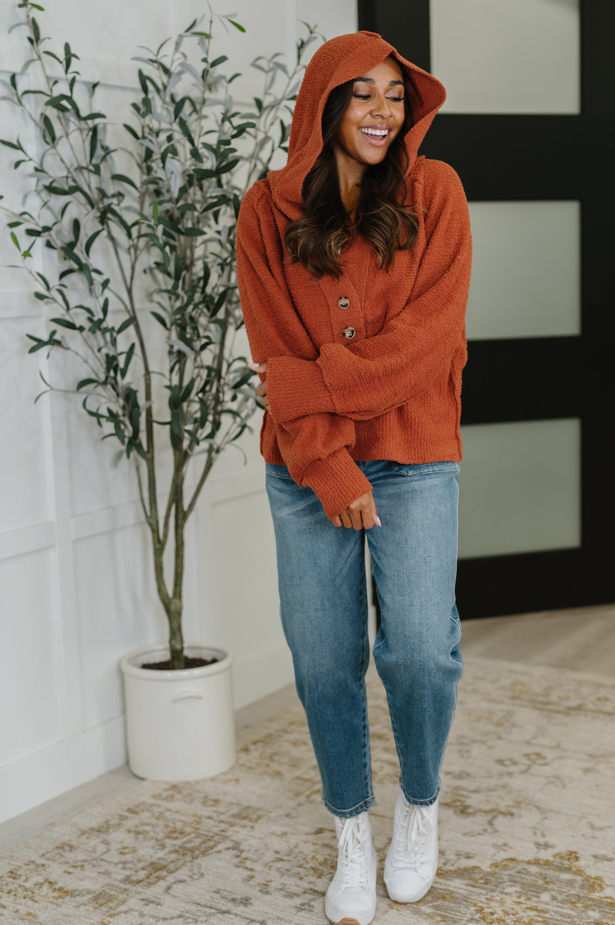 Woman wearing a rust-colored hoodie and blue jeans standing indoors next to a plant.