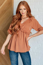 Woman wearing a rust-colored blouse and blue jeans against a white brick wall.