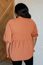 Person wearing a rust-colored blouse against a white brick wall.