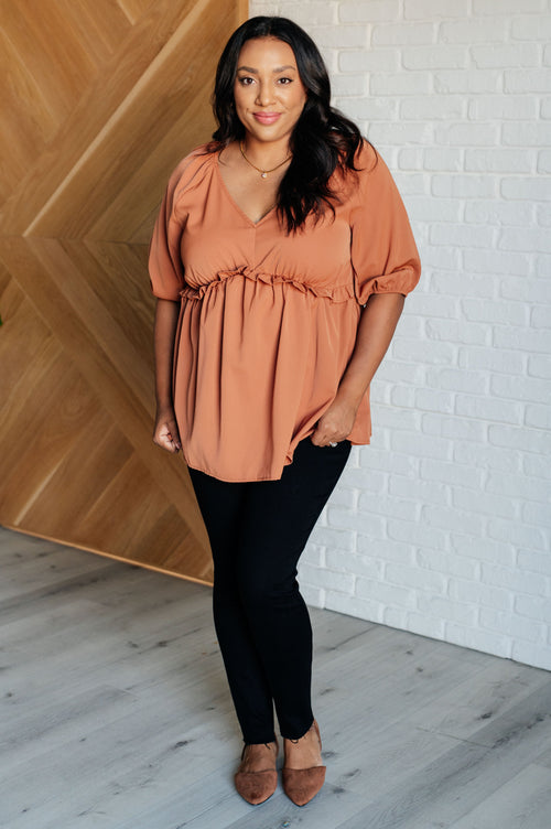 Woman wearing a rust-colored blouse and black pants standing against a white brick wall.