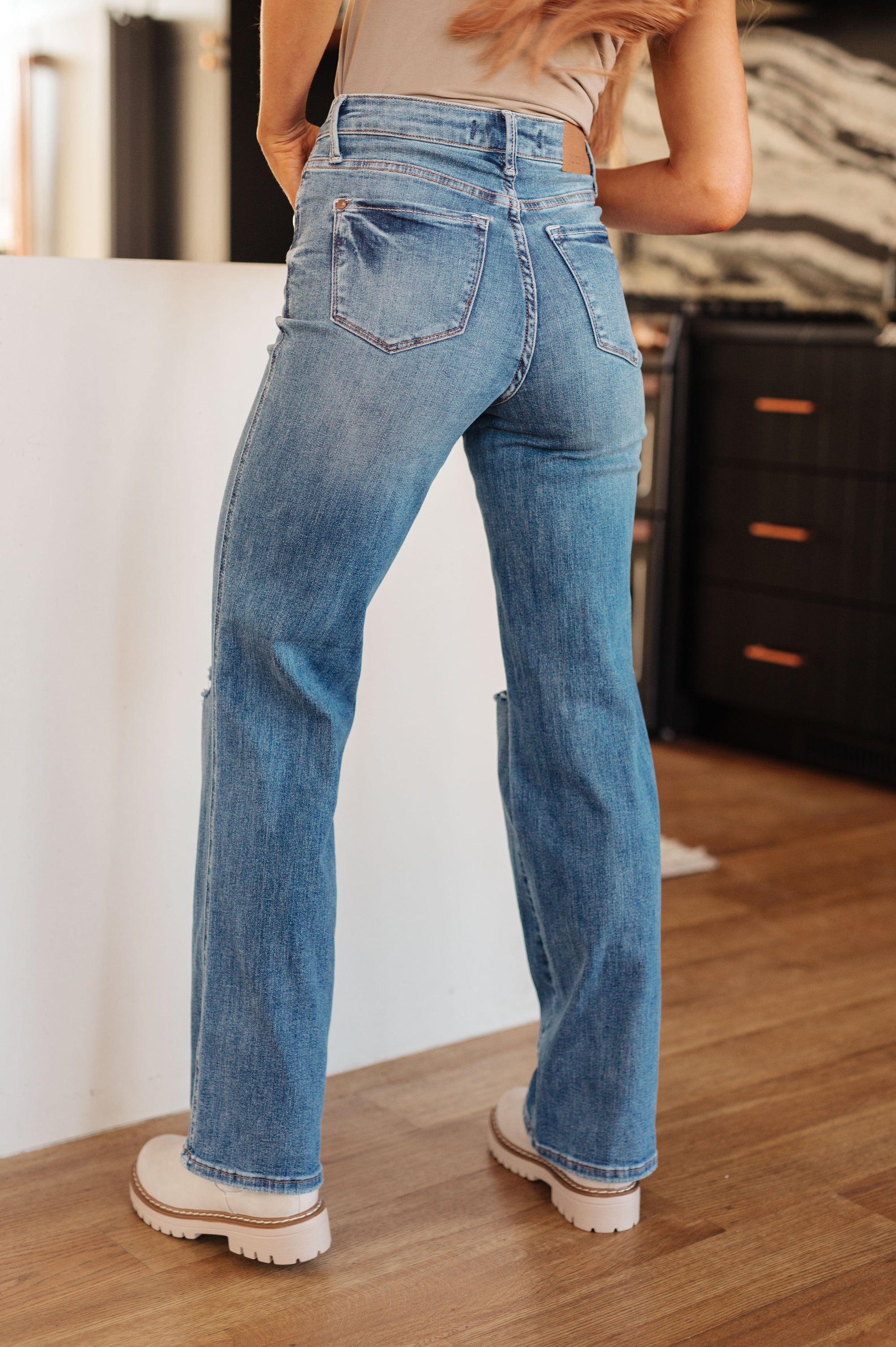 Person wearing blue jeans standing in a room with a white wall and wooden floor.