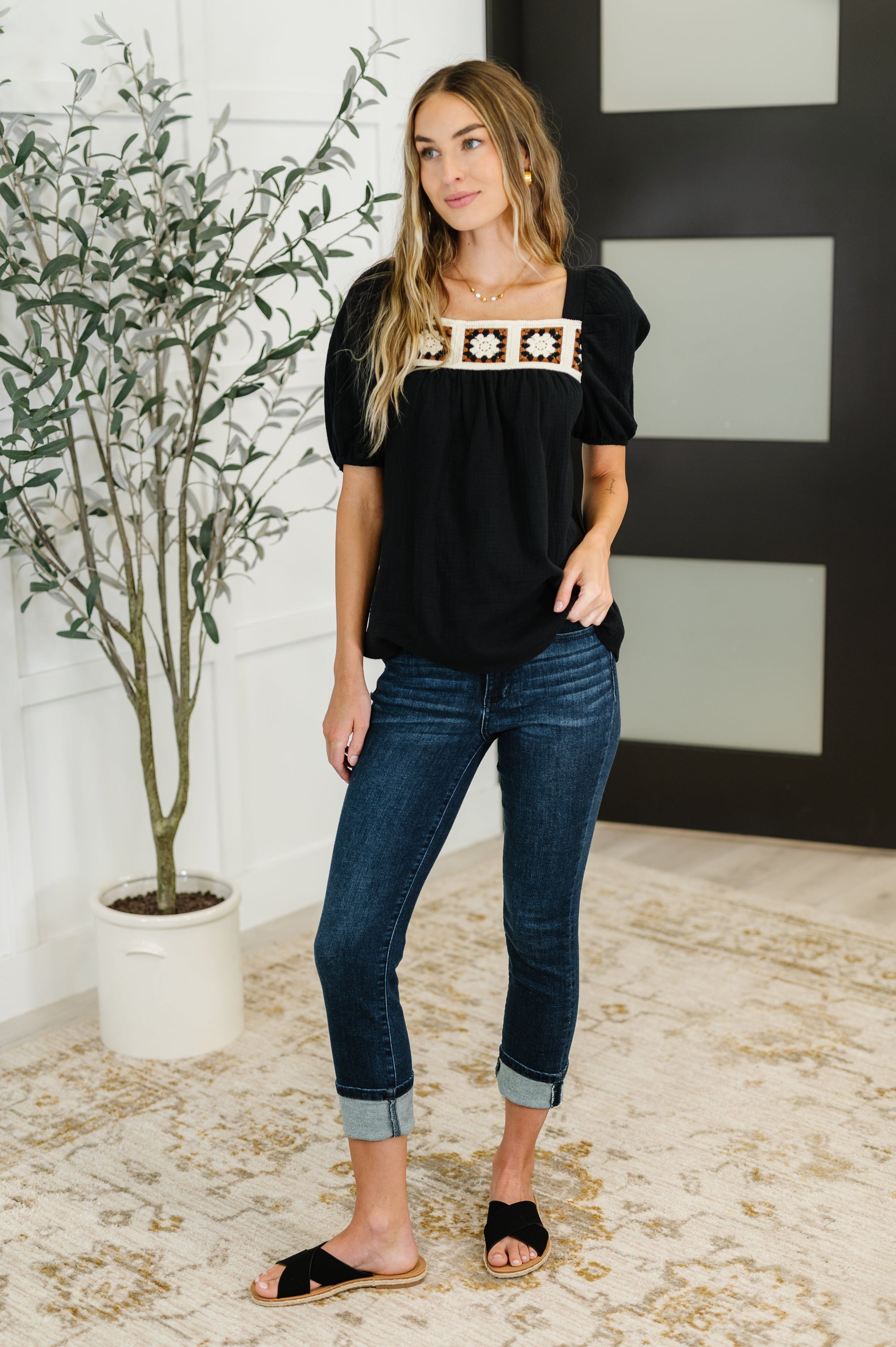 Woman wearing a black top with a patterned neckline and blue jeans standing indoors.