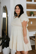 Woman wearing a white dress standing in a room with a mirror and shelves.