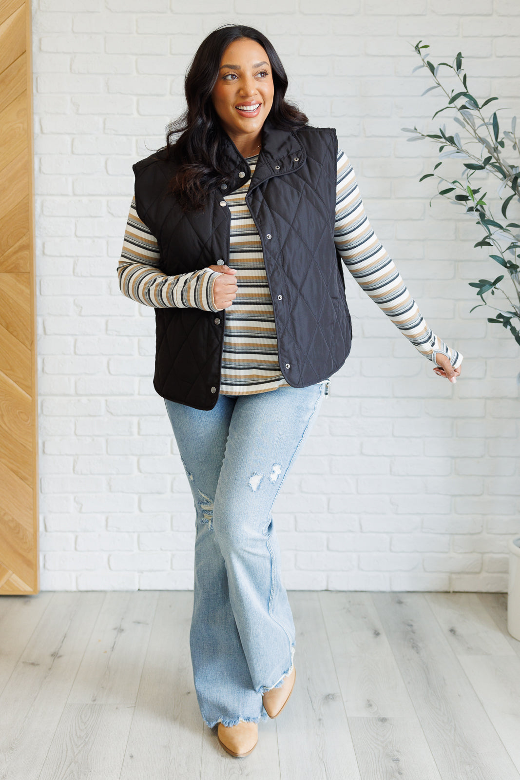 Woman wearing a black quilted vest over a striped shirt with light blue jeans indoors.