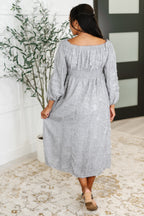 Woman wearing a gray dress standing in a room with a plant and door in the background.