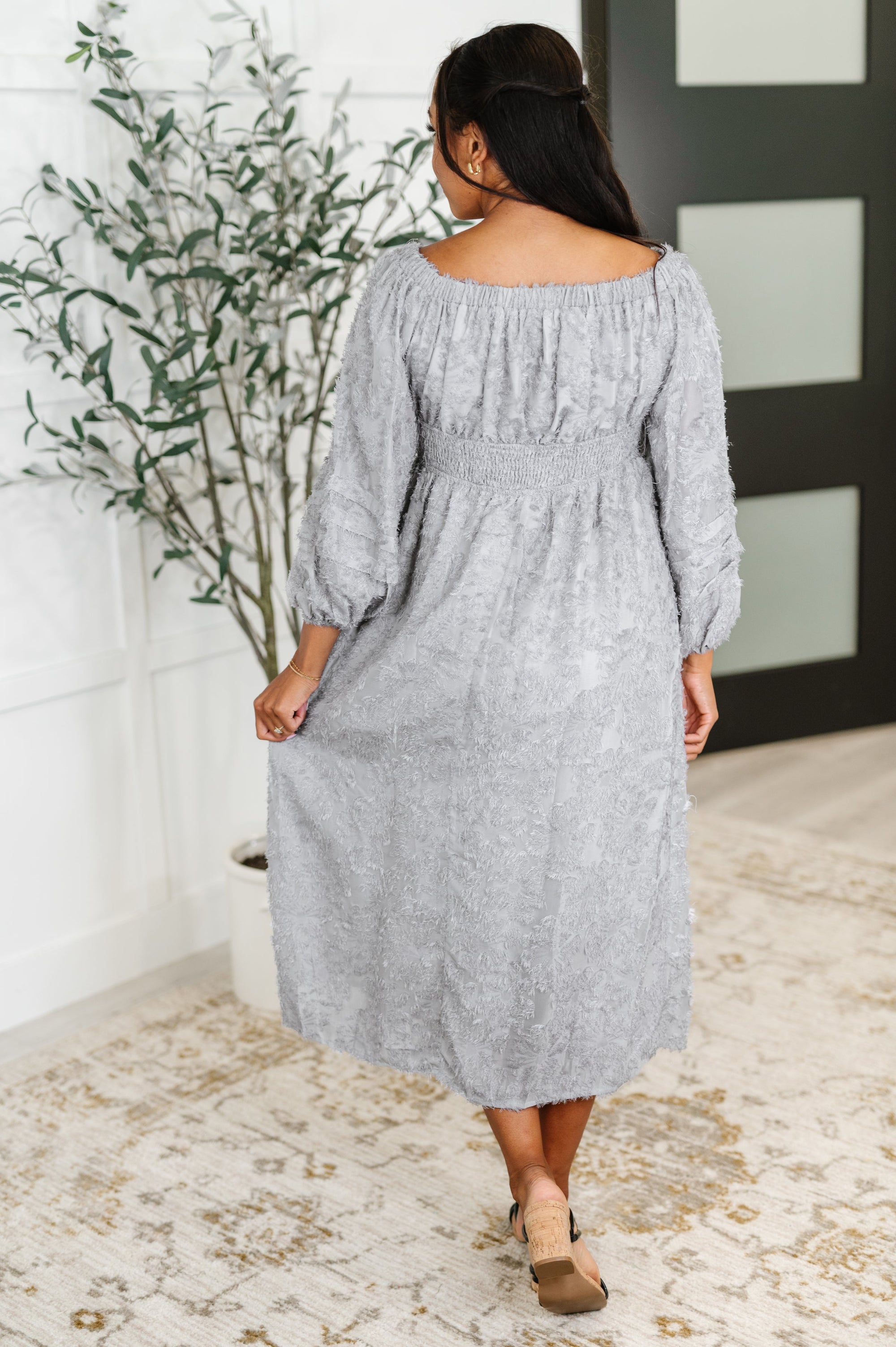 Woman wearing a gray dress standing in a room with a plant and door in the background.
