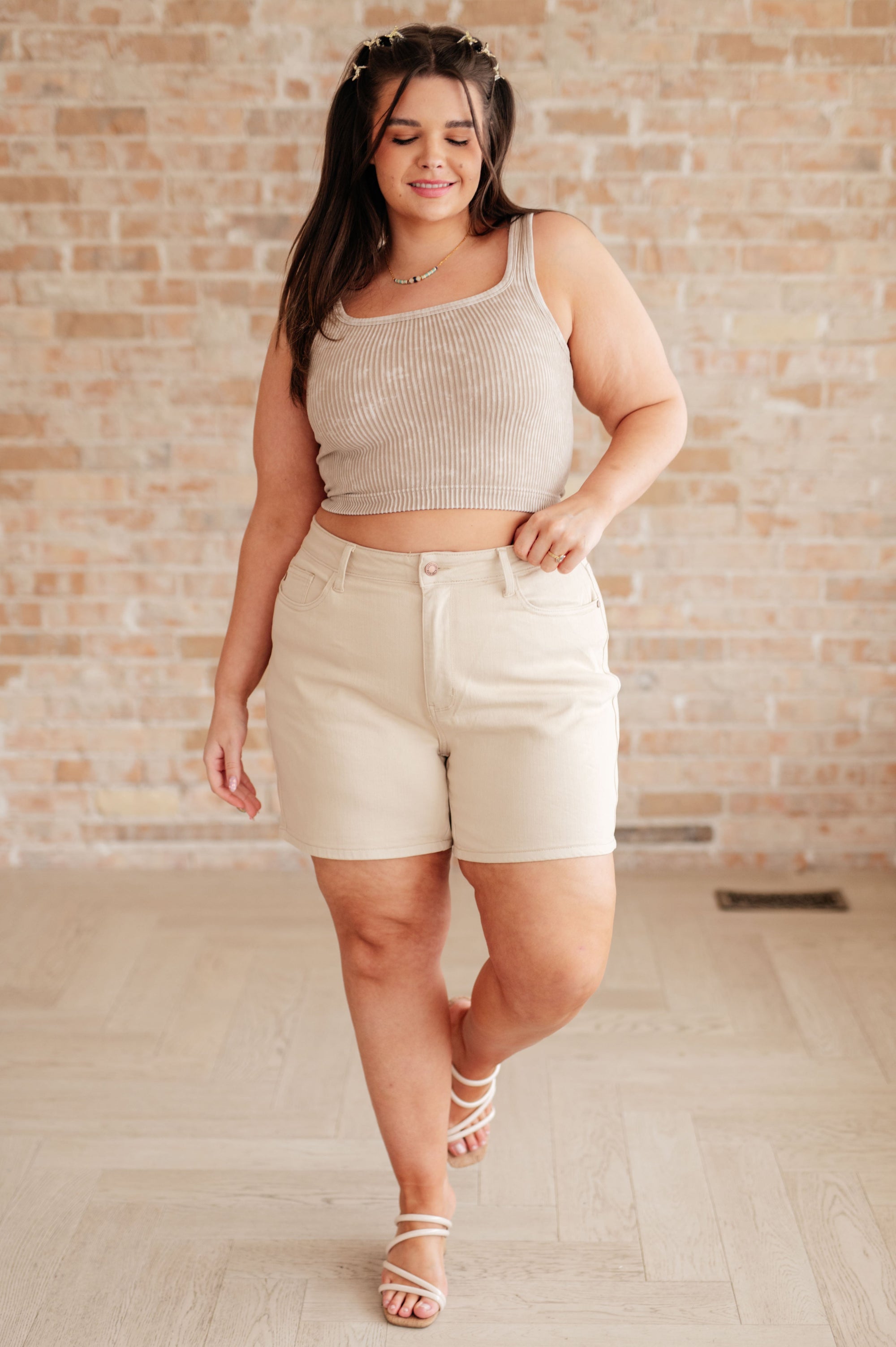 Woman wearing a beige tank top and shorts against a brick wall.
