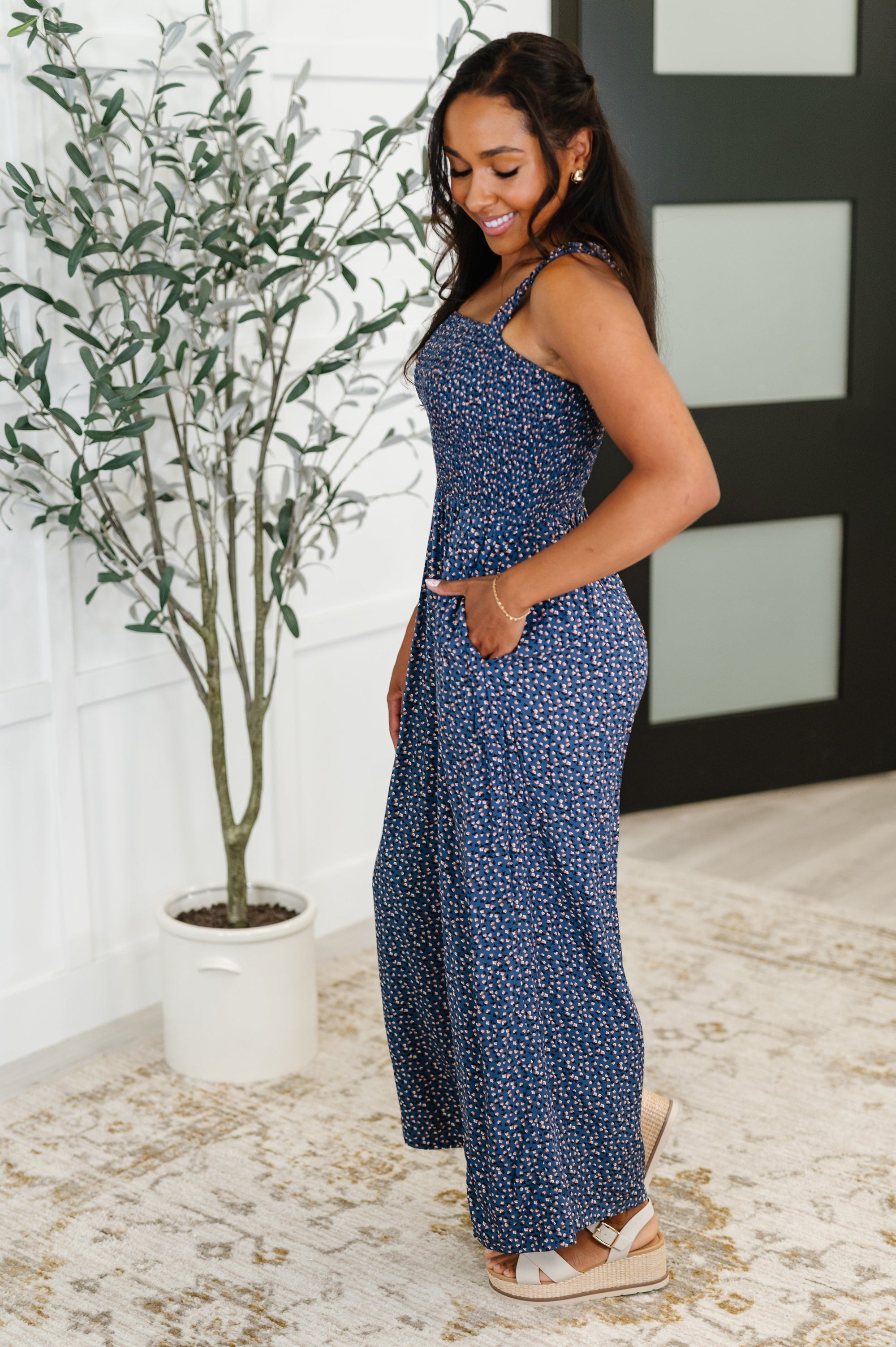 Woman wearing a blue floral jumpsuit standing next to a potted plant indoors.