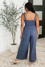 Woman wearing a blue floral jumpsuit standing in a room with a plant and white wall.