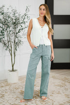 Woman wearing a white sleeveless top and light blue pants standing indoors with a plant in the background.