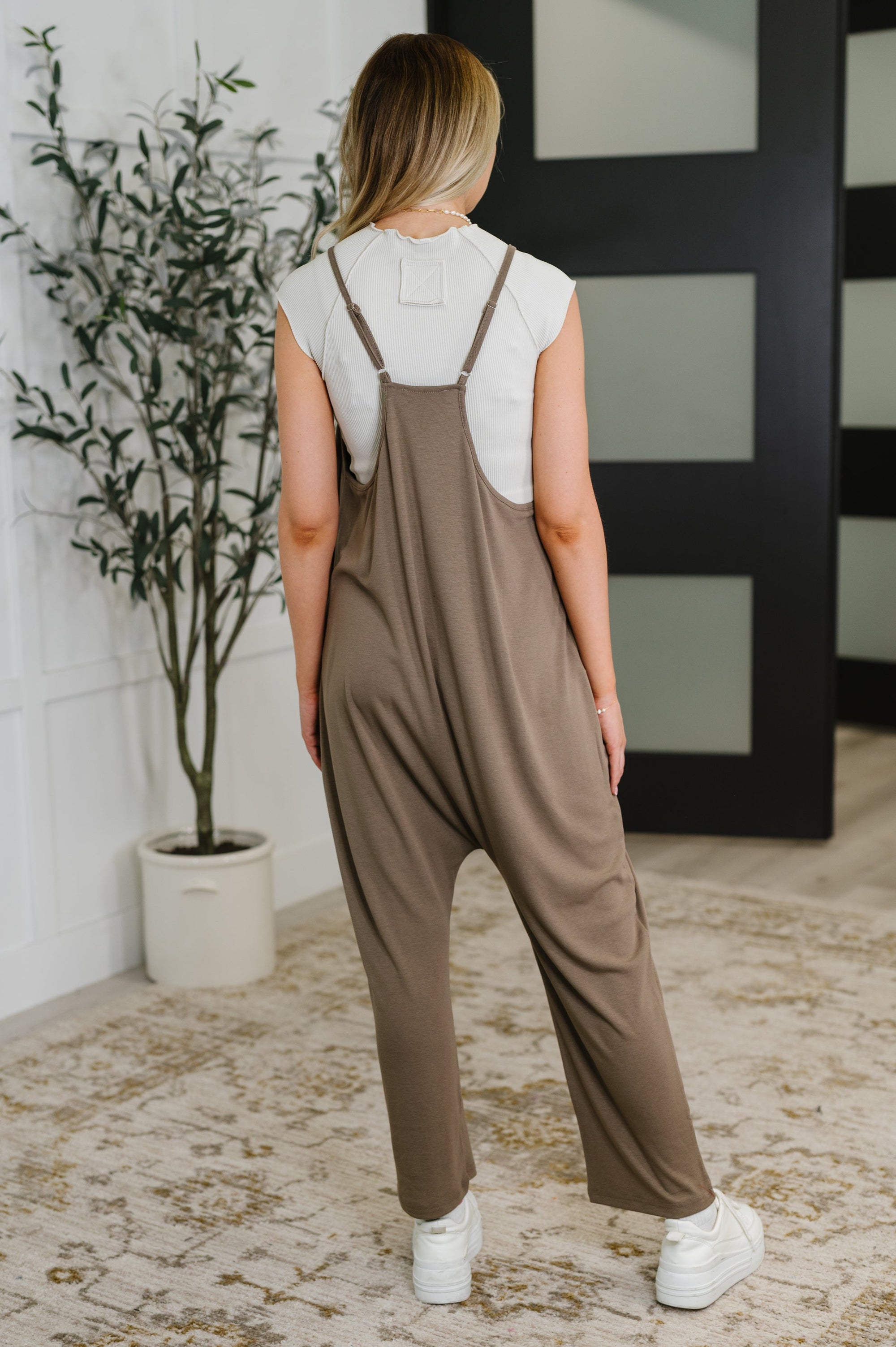 Person wearing a brown jumpsuit standing in a room with a plant and decorative wall.