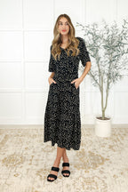 Woman wearing a black floral dress standing in a room with a white tiled wall and a plant.