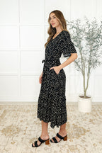 Woman wearing a black floral dress standing in a room with a plant and tiled floor.