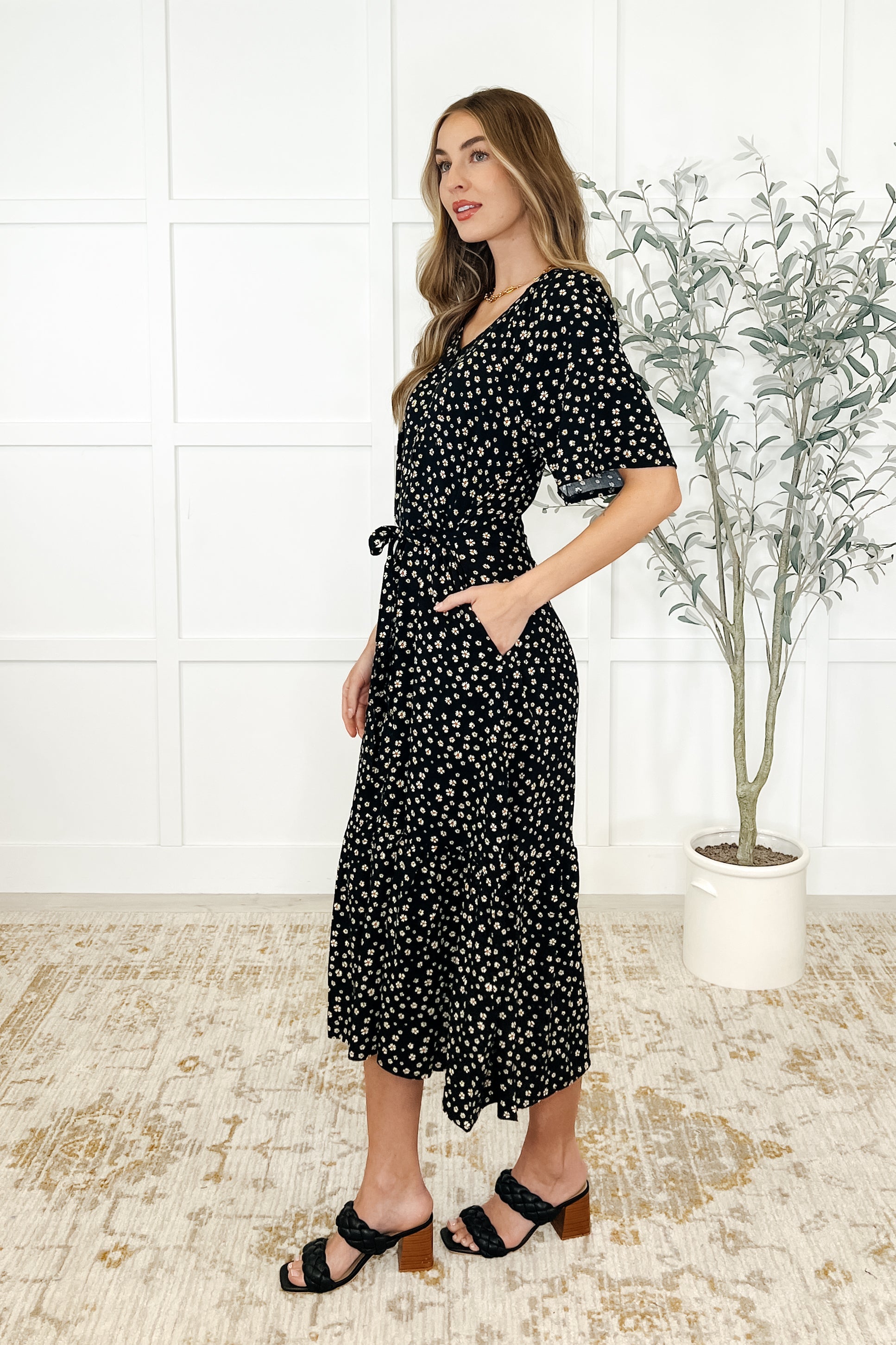 Woman wearing a black floral dress standing in a room with a plant and tiled floor.