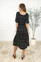 Woman wearing a black floral dress standing in a room with a white wall and plant.