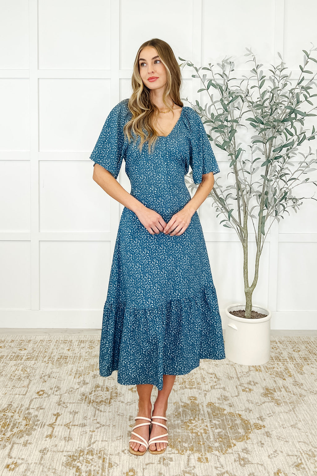 Woman wearing a blue floral dress standing in a room with a white wall and a plant.