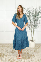Woman wearing a blue floral dress standing in a room with a white wall and a plant.