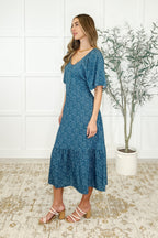 Woman wearing a blue dress standing in a room with a plant and white walls.