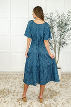 Woman wearing a blue floral dress standing in a room with a white wall and plant.