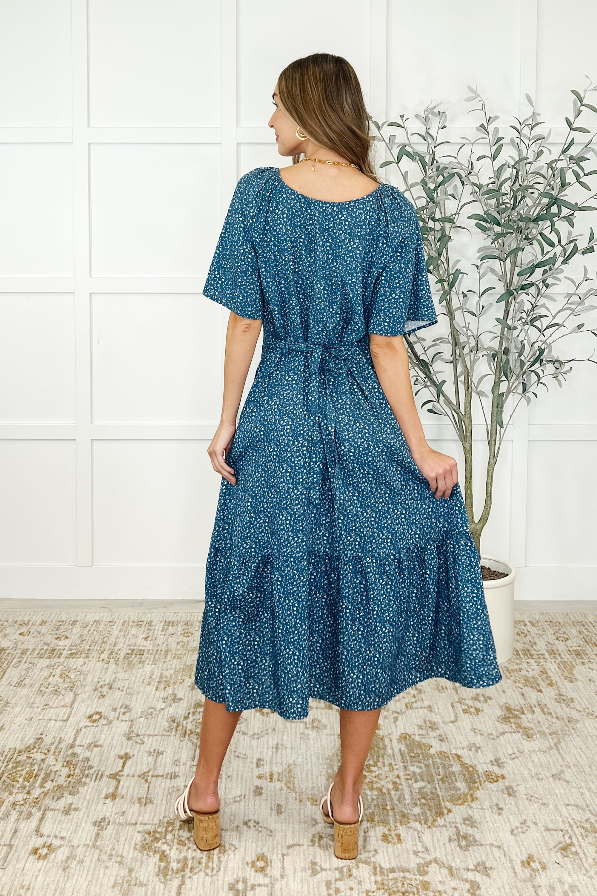Woman wearing a blue floral dress standing in a room with a white wall and plant.