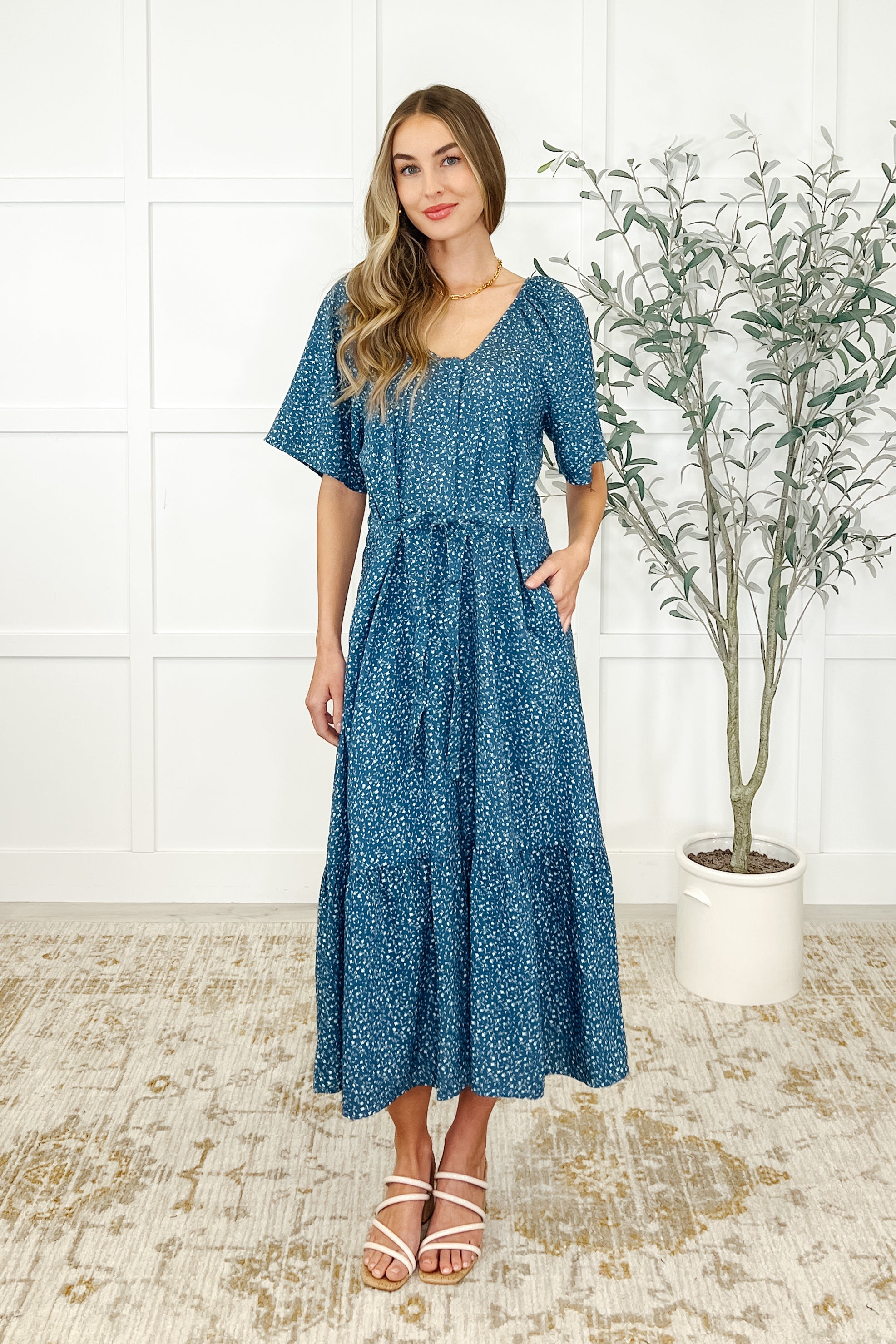 Woman wearing a blue patterned dress standing in a room with a white wall and a plant.
