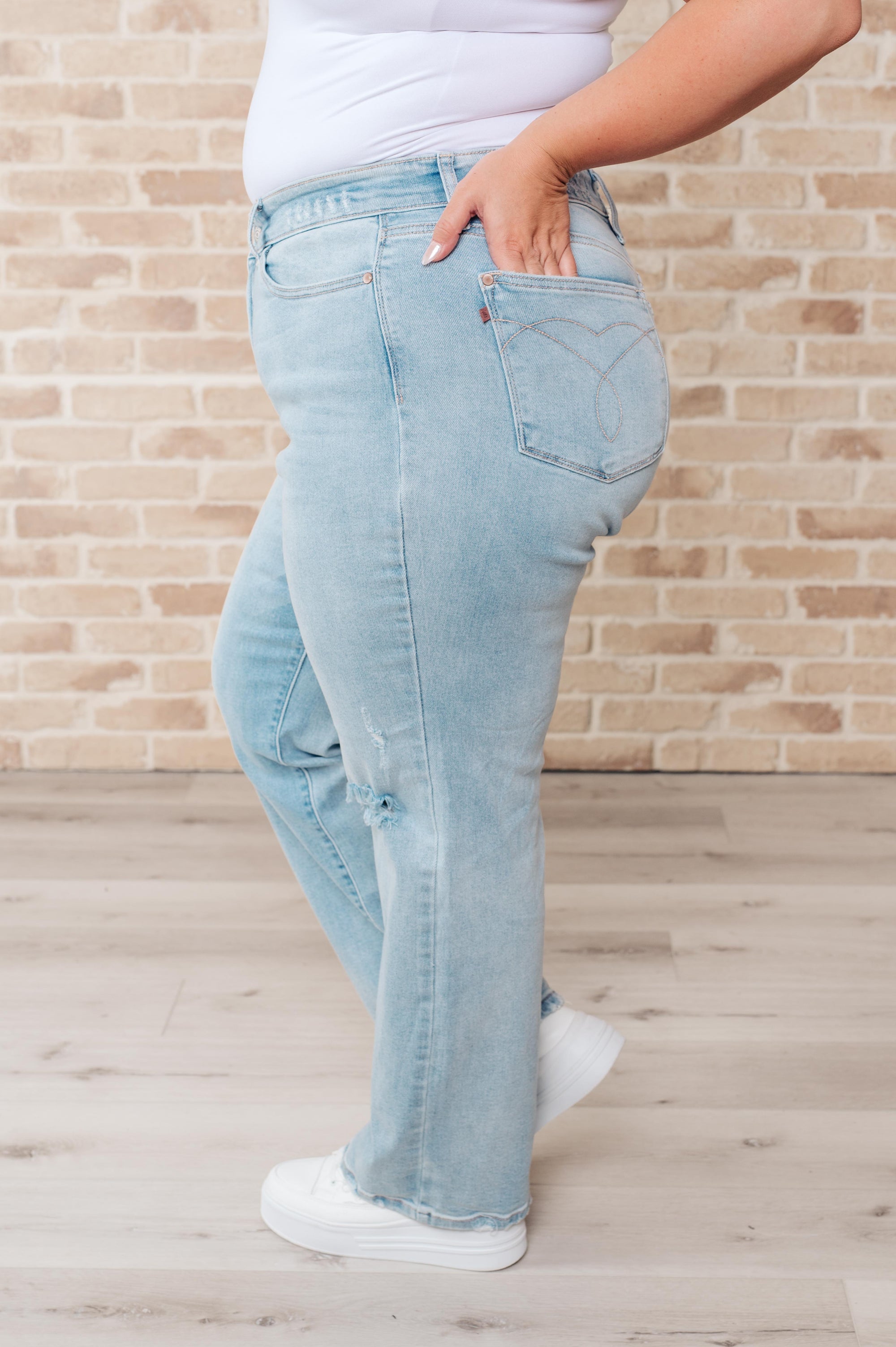 Person wearing light blue jeans and white sneakers against a brick wall.