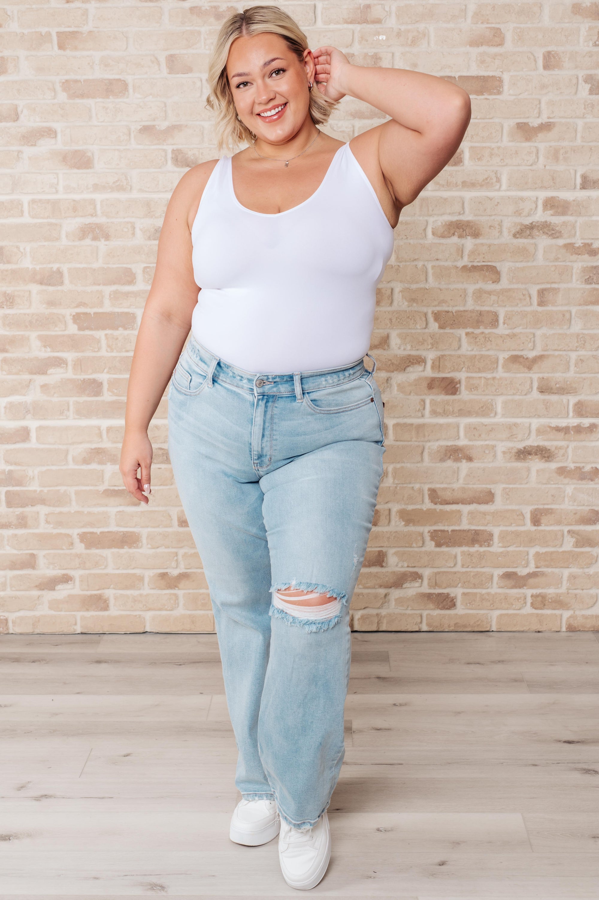 Woman wearing a white tank top and light blue jeans against a brick wall.