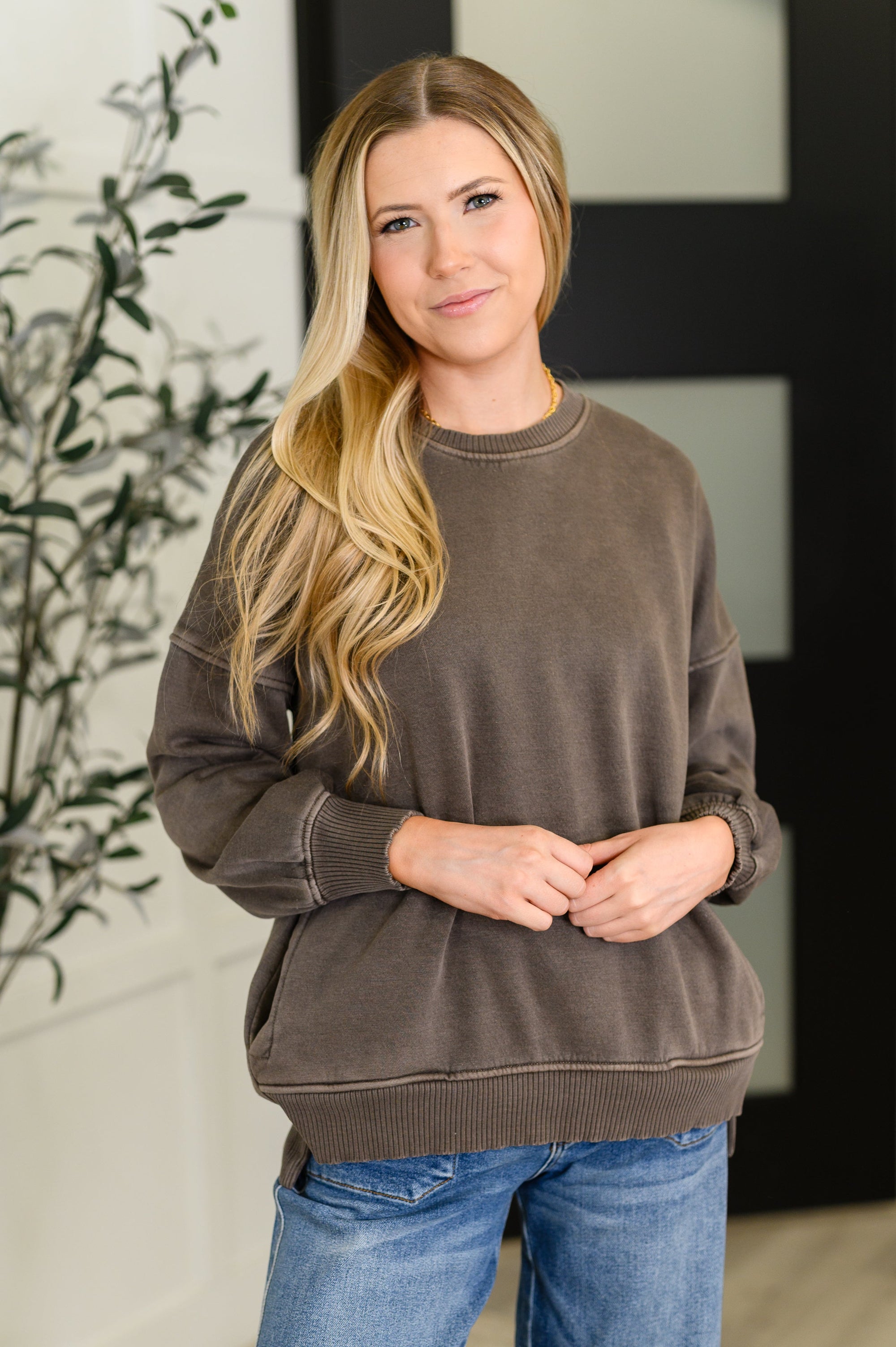 Woman wearing a brown sweater and blue jeans standing indoors with a blurred background