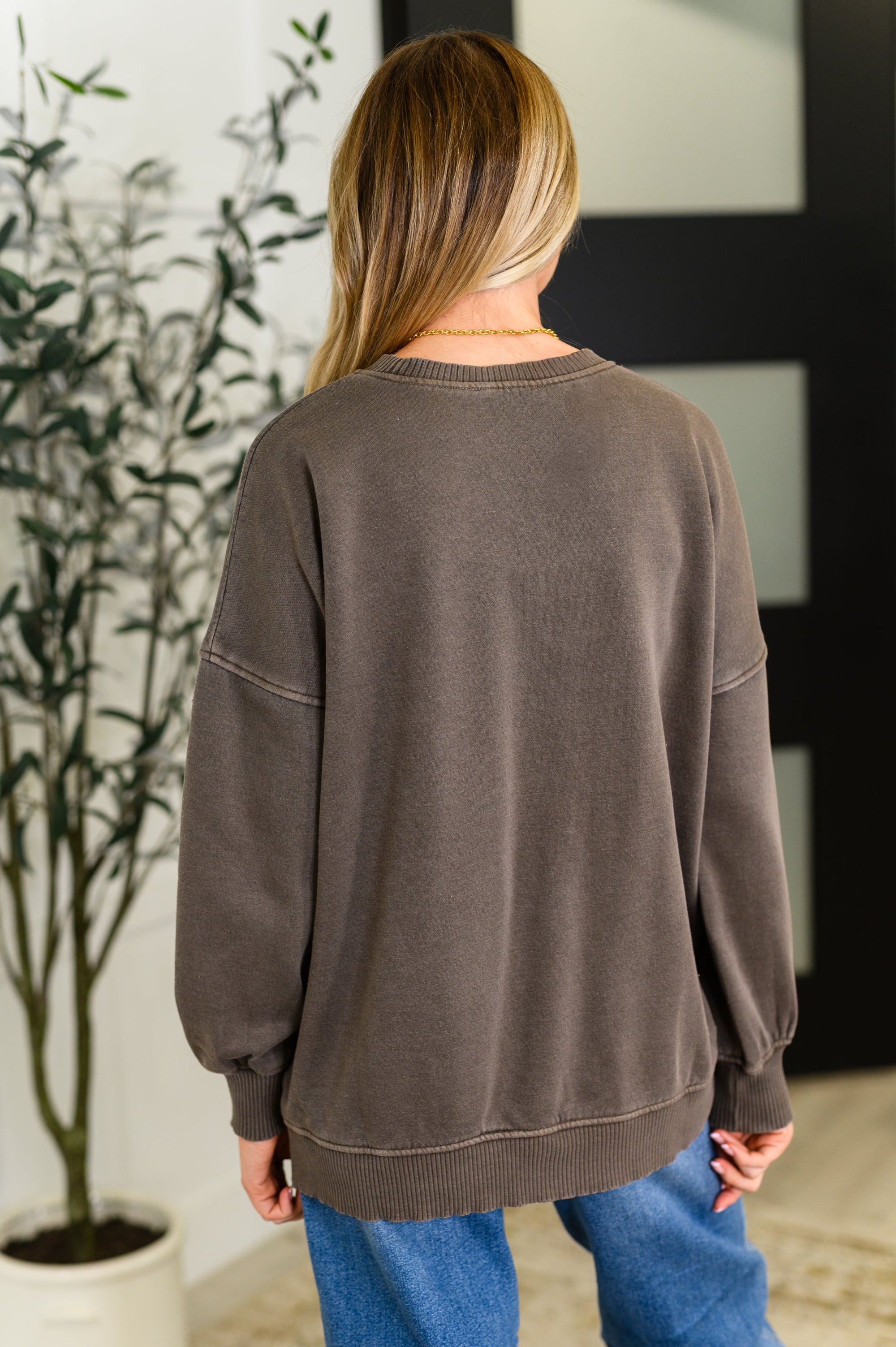 Person wearing a brown sweatshirt and blue jeans standing in a room with a plant and framed picture.