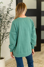Person wearing a green sweatshirt in an indoor setting with a plant and mirror in the background.