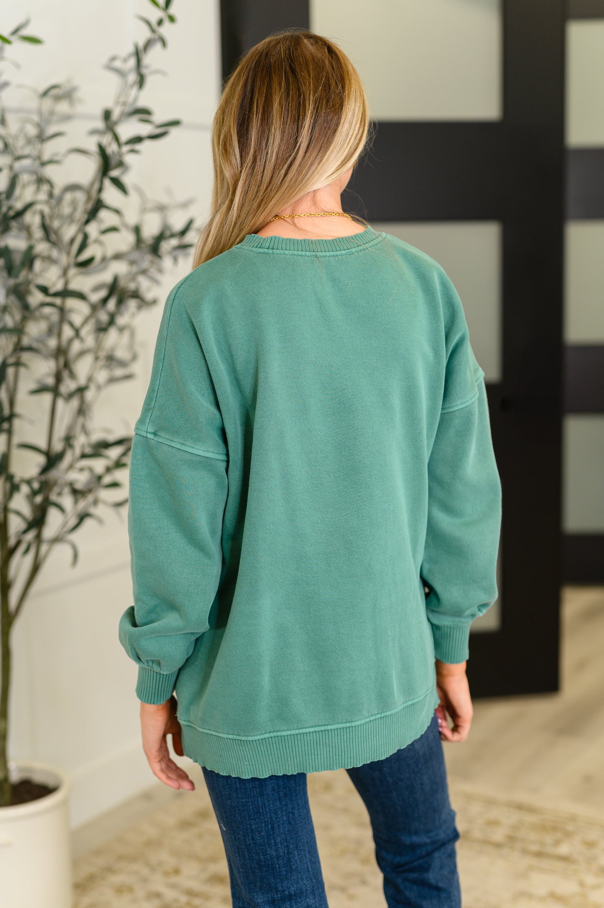 Person wearing a green sweatshirt in an indoor setting with a plant and mirror in the background.