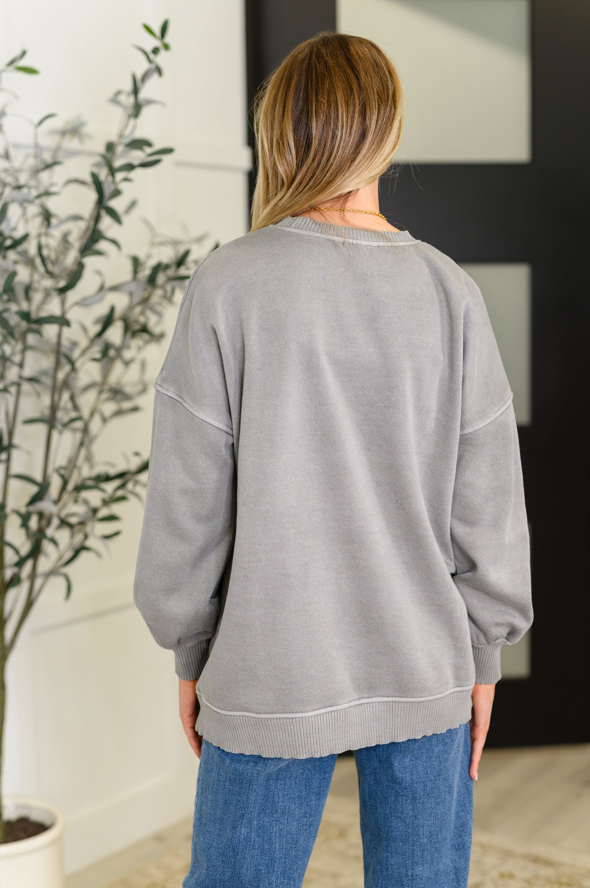 Person wearing a gray sweatshirt and blue jeans standing in a room with a plant and framed picture.