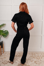 Person wearing a black jumpsuit standing in a room with a white wall and plant.