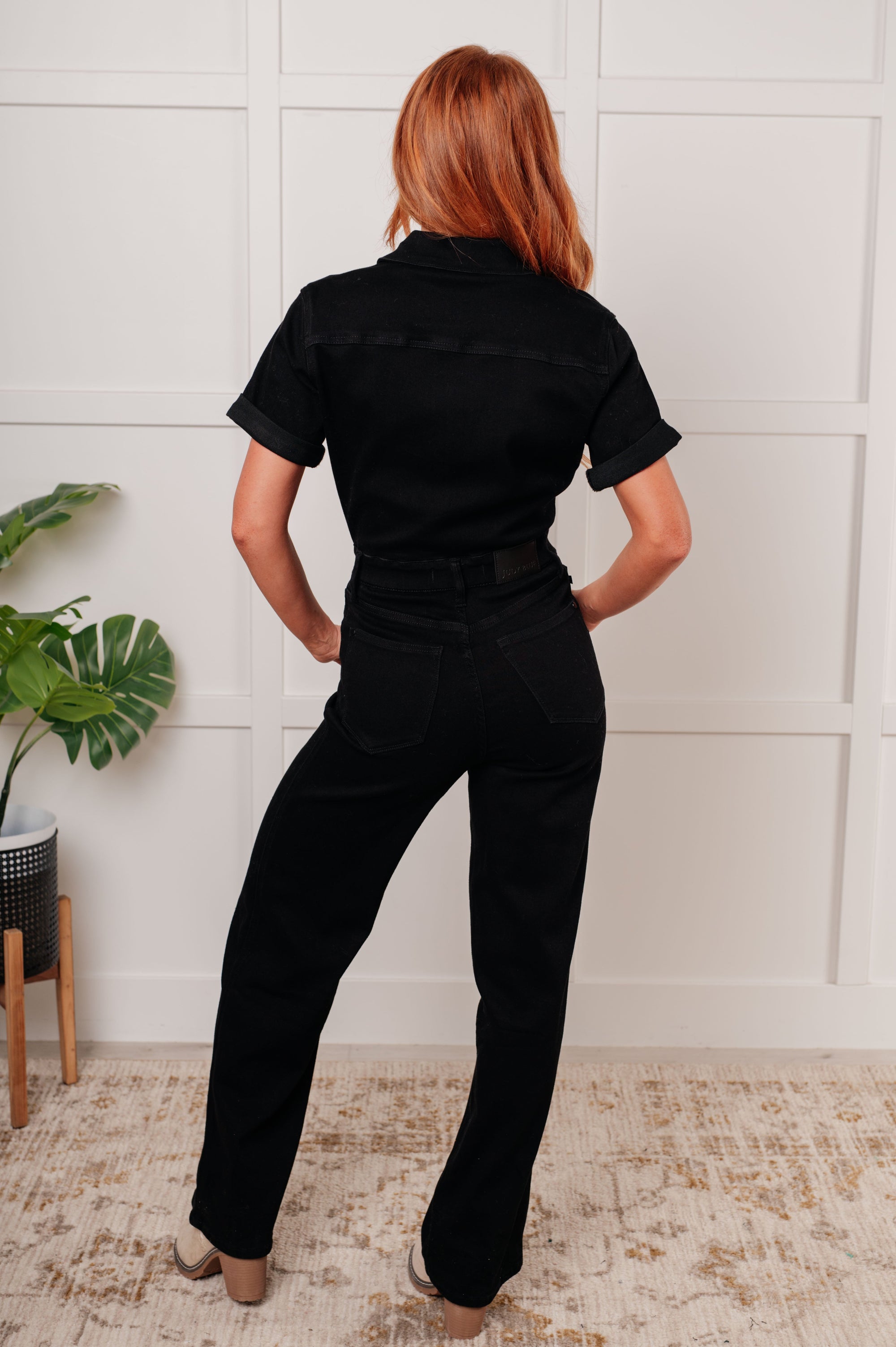 Person wearing a black jumpsuit standing in a room with a white wall and plant.