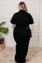 Person wearing a black outfit standing in a room with a plant and wooden furniture.
