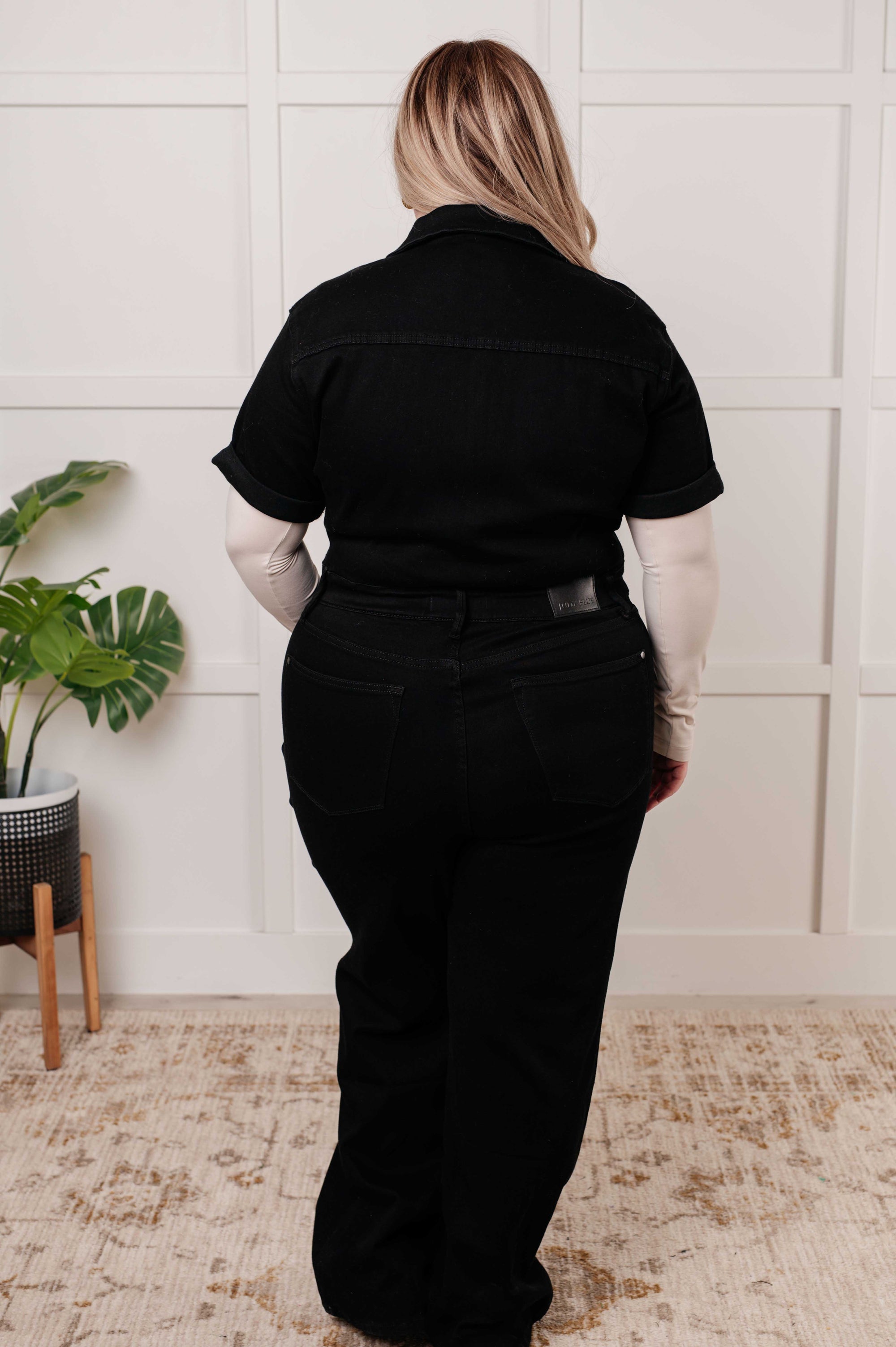 Person wearing a black outfit standing in a room with a plant and wooden furniture.