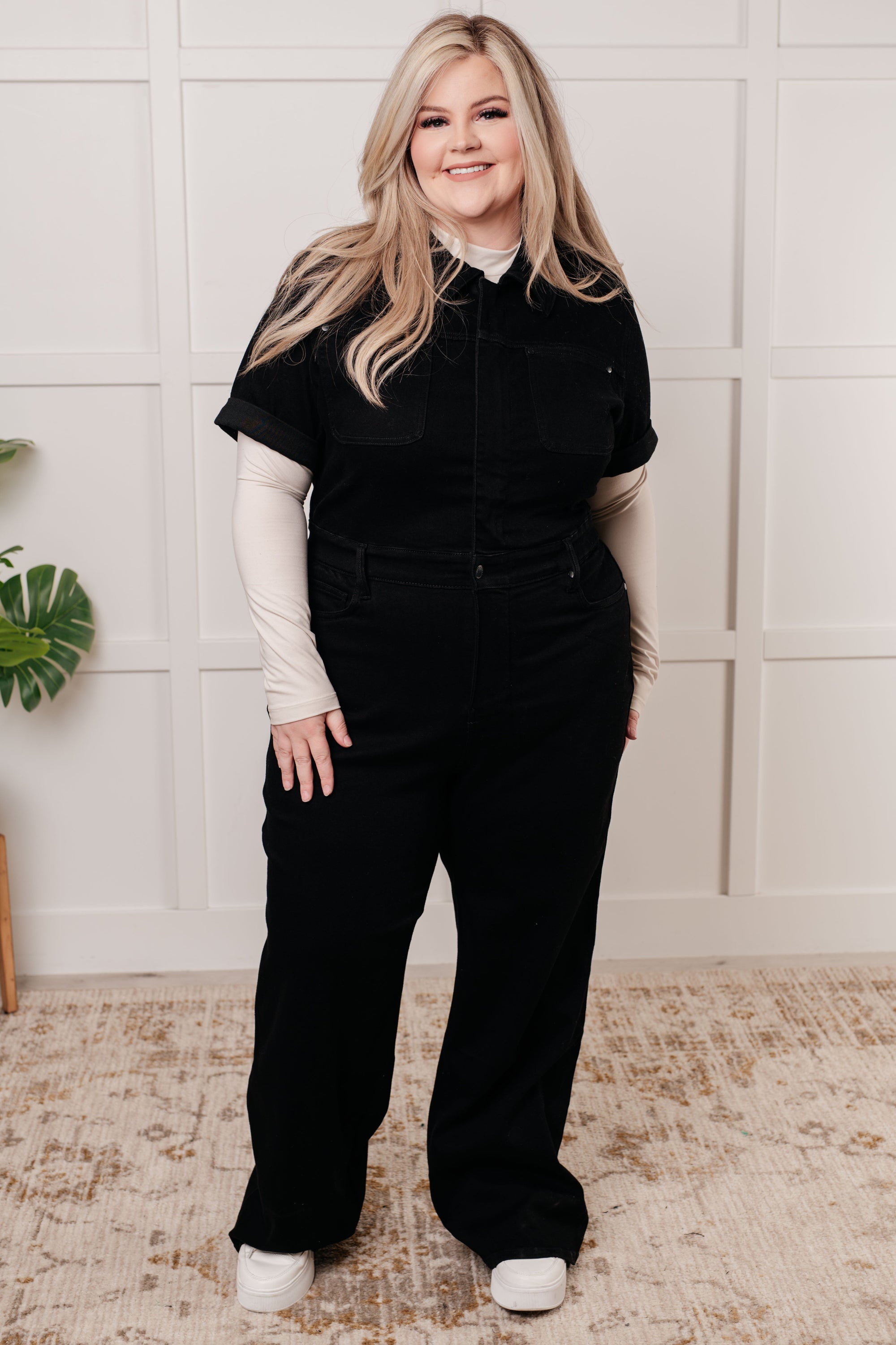 Woman wearing a black jumpsuit with a white shirt underneath, standing against a light-colored wall.