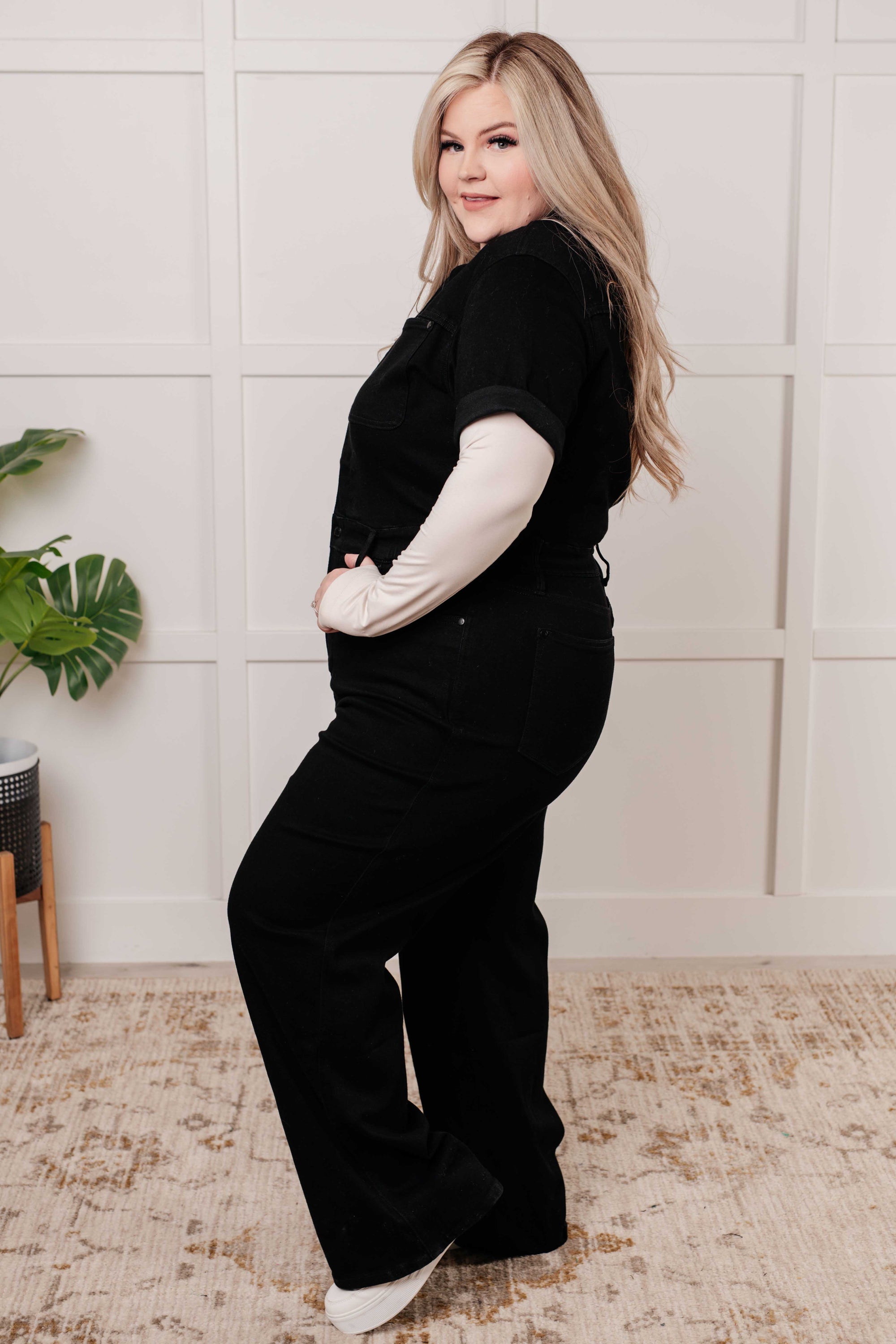 Woman wearing a black outfit with white sleeves in a room with a plant and rug.