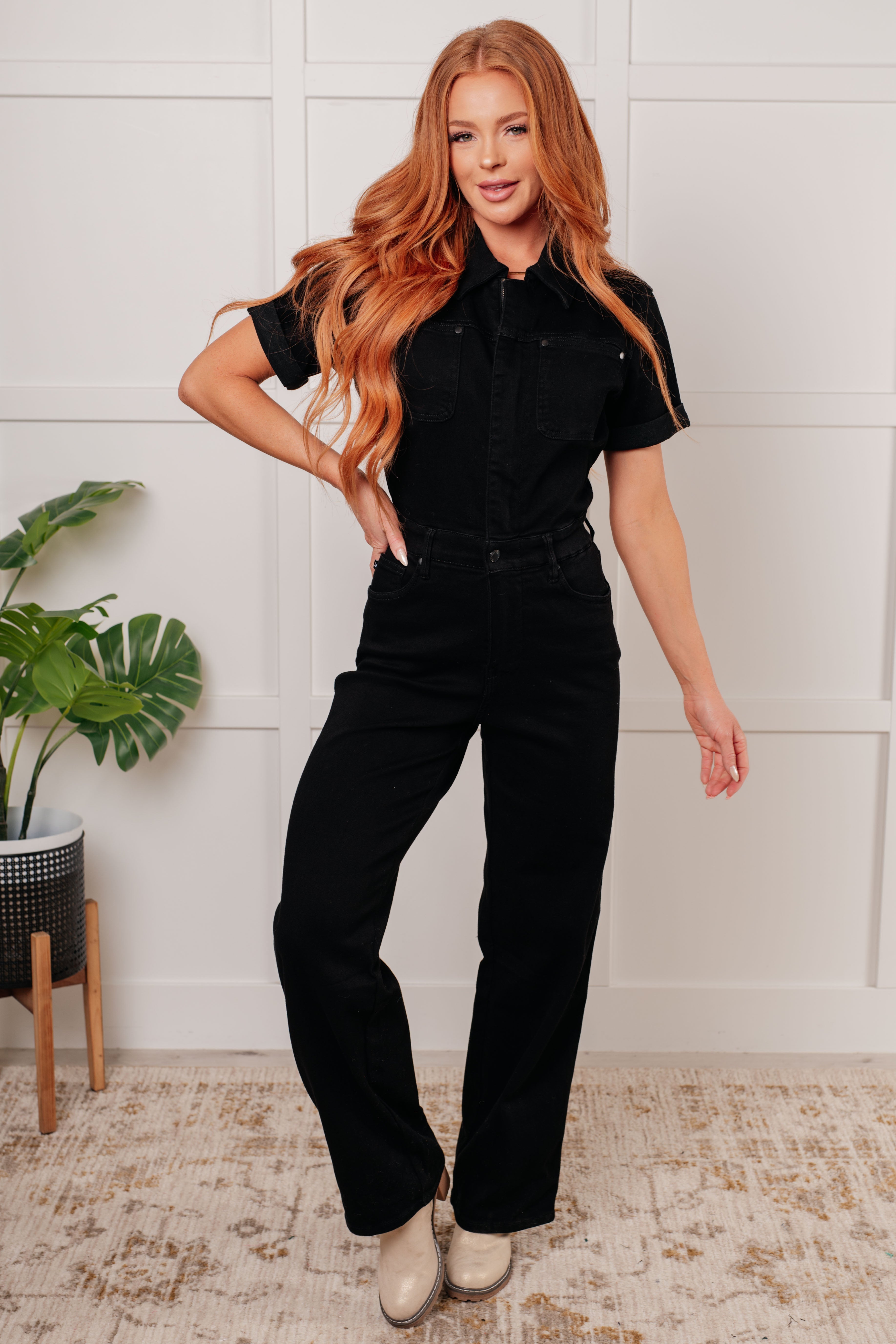 Woman wearing a black outfit standing in a room with a plant and white wall.