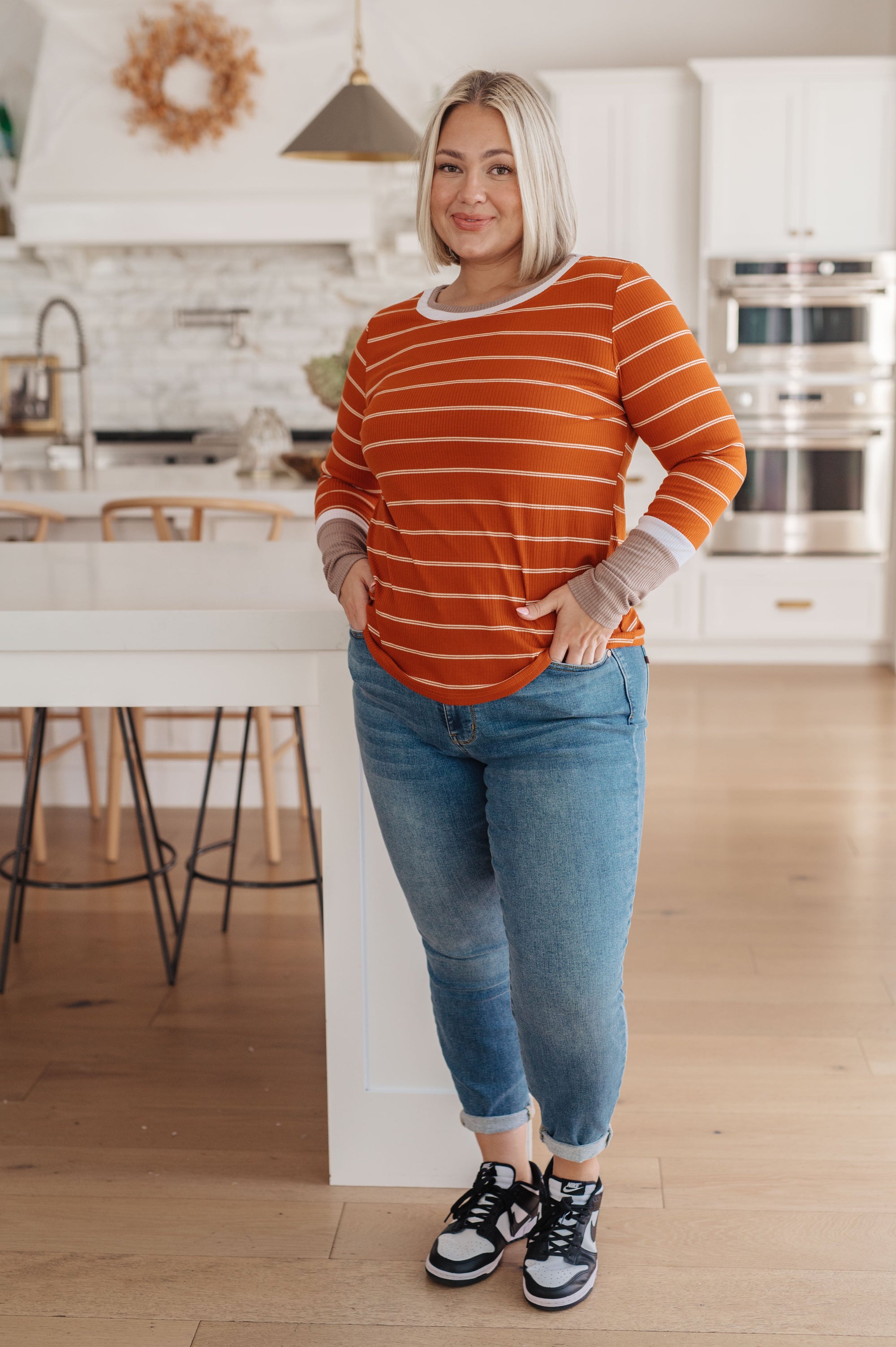 Woman wearing an orange striped sweater and blue jeans standing in a kitchen.