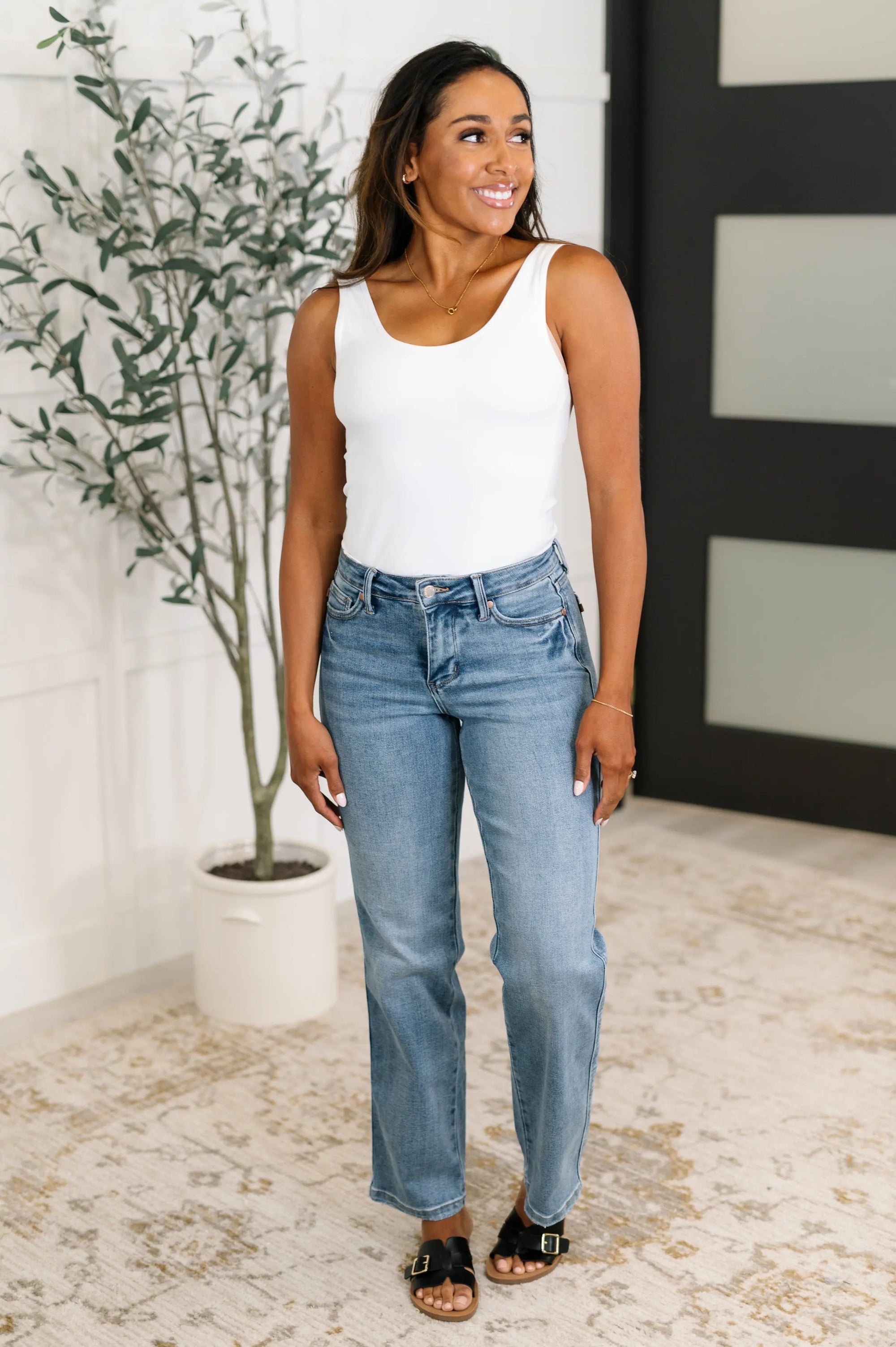 Woman wearing a white tank top and blue jeans standing indoors with a plant in the background.